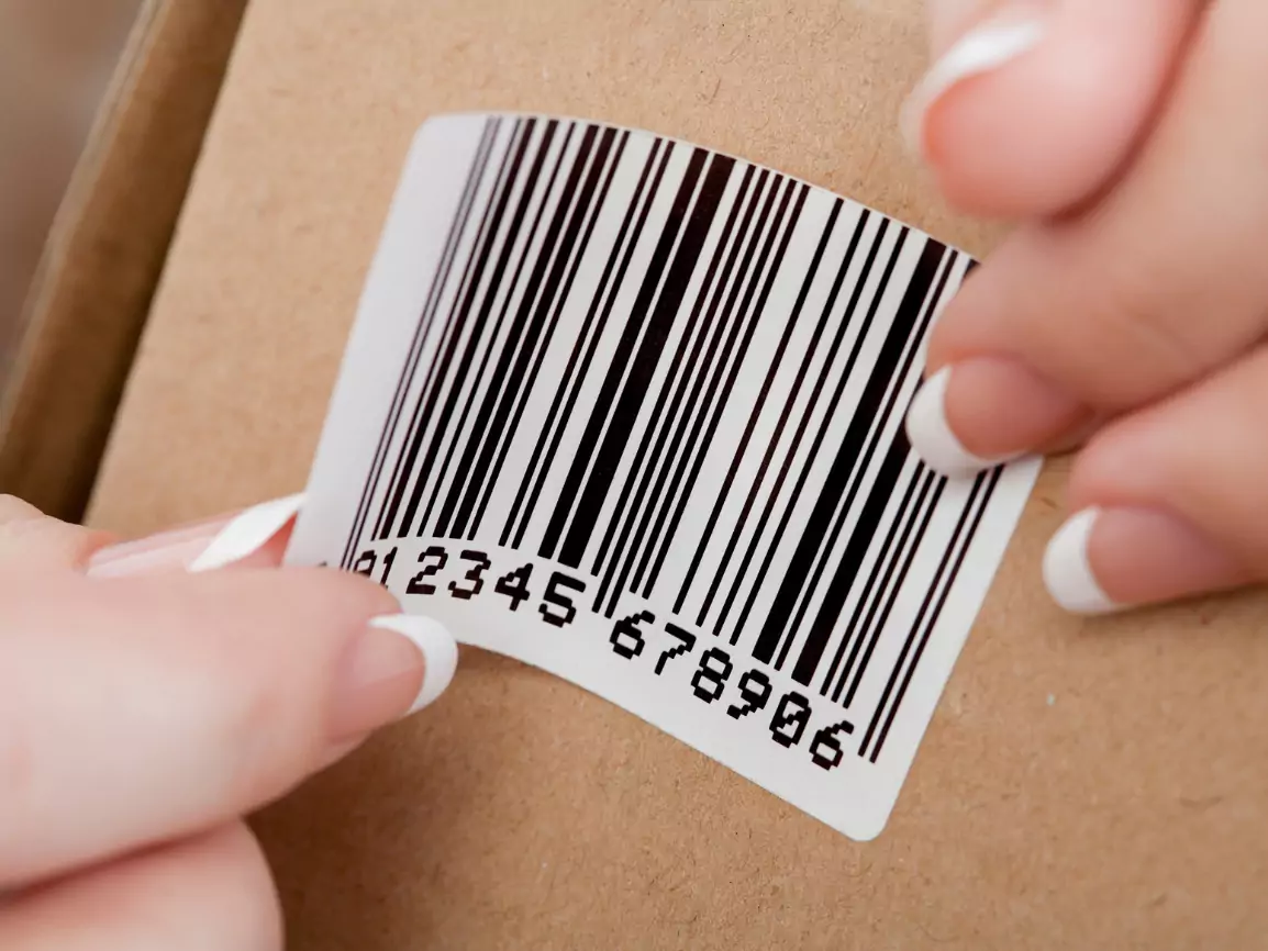 A person is applying a barcode label onto a cardboard surface with their fingers.